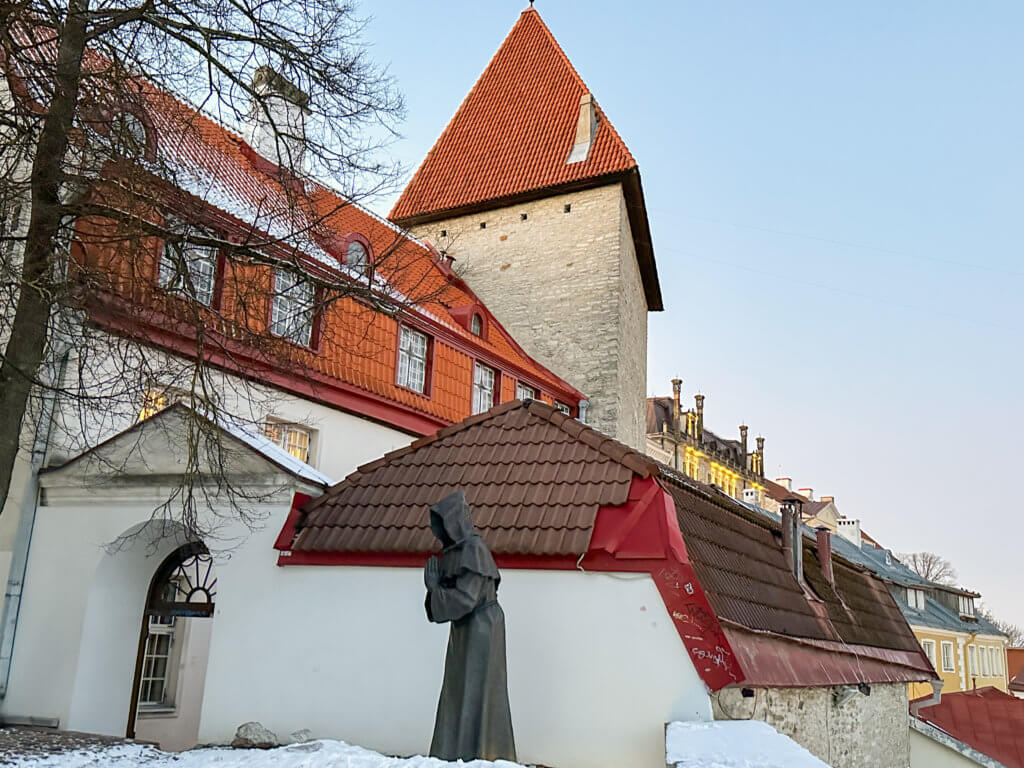 Danish Kings Garden in Tallinn Estonia. A medium-angle, eye-level shot captures a bronze statue of a hooded, faceless monk in a praying stance, standing in front of a white building with a brown tiled roof in Tallinn, Estonia. The statue, positioned in the center, is made of dark bronze and has a hooded cloak that obscures its face. The building behind it has a white facade and a brown tiled roof with a red trim. To the left, a large, leafless tree stands in front of another white building with a red tiled roof and several windows. In the background, a tall, square stone tower with a red conical roof rises above the other buildings. The ground is covered in snow, and the sky is a clear, light blue. The lighting is bright and even, suggesting a clear day.