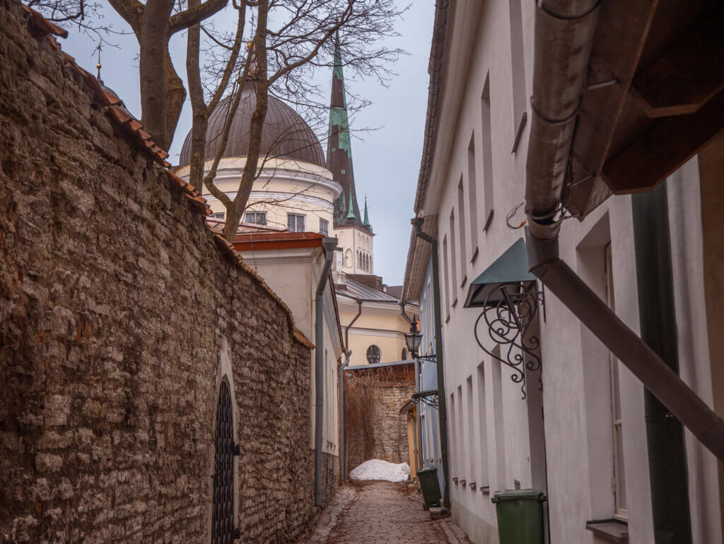 A vertical, eye-level shot looking down a narrow, damp cobblestone alleyway in Tallinn, Estonia. On the left, a tall medieval wall made of rough, grey limestone stretches into the distance, featuring a dark wrought-iron arched gate. On the right, a white building with a dark metal downspout and decorative wrought-iron window awnings lines the path. In the background, the dark, rounded dome of a church and a tall, slender green spire rise above the rooftops against a pale, overcast sky. A small pile of white snow is visible at the far end of the alley.