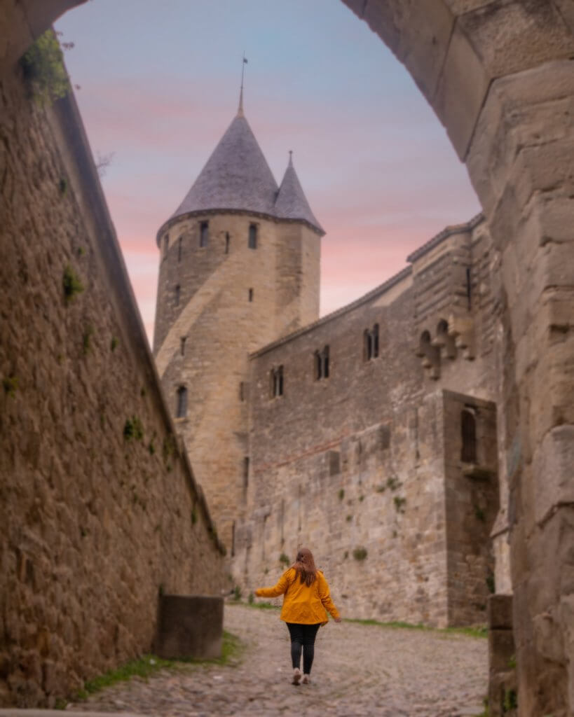 A medium-angle shot captures a person with long reddish-brown hair, wearing a yellow jacket and black pants, walking away from the camera up a narrow, cobblestone path in Cite de Carcassonne, France.

The path is framed by massive, historic stone walls that lead toward a prominent fortified tower with a tall, conical grey roof. A portion of a stone archway frames the top right of the scene, and the background features a soft pink and blue sunset sky.