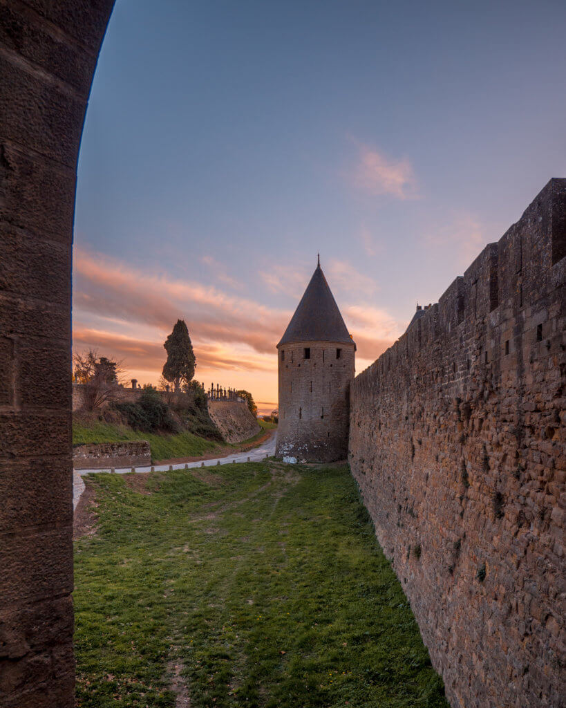 Fairytale turrets of the city walls of Carcassonne France. A wide-angle, eye-level shot captures a dramatic view along the towering outer fortifications of Carcassonne, France.

On the right, a massive stone defensive wall extends deep into the background, leading to a tall, circular fortified tower topped with a dark, conical slate roof. To the left of the wall, a grassy moat area runs alongside a paved path. In the distance, silhouettes of trees and a hillside are visible against a vibrant sunset sky filled with soft orange, pink, and purple clouds. The scene is framed on the far left by the dark, textured edge of another stone structure in the immediate foreground.