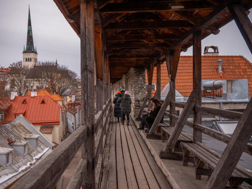 People walking on the wooden walkway of Hellemann Tower in Old Town Tallinn. A portrait-oriented view from an elevated wooden walkway along a medieval stone wall in Tallinn, Estonia. The walkway features thick wooden support beams and a railed gallery where several people are walking or sitting on wooden benches. Below the gallery, a narrow cobblestone street winds between historic buildings with steep red and orange-tiled roofs. In the background, the cityscape of the Old Town is visible under a grey, overcast sky, highlighted by the tall, slender white tower and dark green spire of St. Olaf's Church.