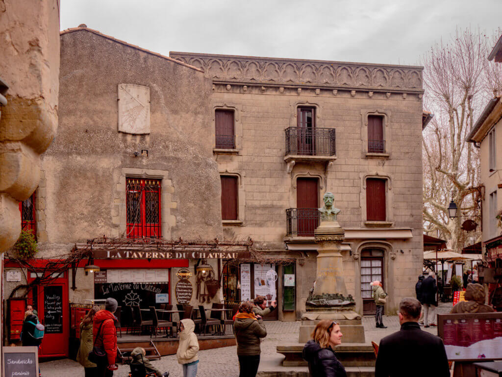 Courtyard in the medieval old town of Carcassonne France. An eye-level, medium shot of a cobblestone square featuring a historic stone building with a restaurant called "La Taverne du Château" on the ground floor.

The building has several brown shutters on the upper windows and a decorative stone trim along the roofline. In the center of the square stands a bronze bust on a tall stone pedestal. Several people in winter clothing are scattered throughout the square, some looking at the restaurant and others walking past. Bare trees and other stone buildings are visible in the background under an overcast sky.