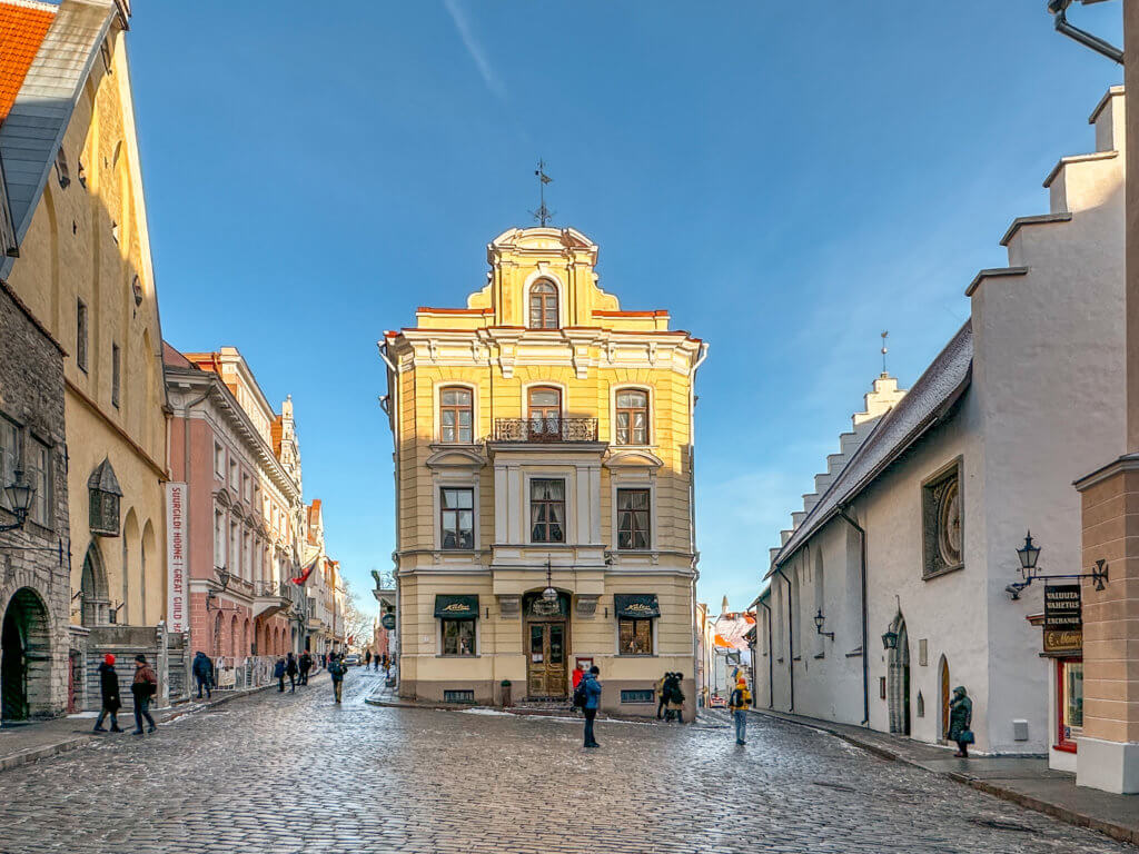 Maiasmokk Cafe the oldest cafe in Tallinn. A vibrant, sunlit view of a narrow cobblestone street fork in Tallinn's Old Town, Estonia. At the center of the fork stands a prominent three-story, pale yellow building with ornate white architectural detailing, a small central balcony, and a decorative roofline with a weather vane. To the left, a street lined with historic multi-story buildings stretches into the distance, including the Great Guild Hall. To the right, a long, white medieval building with a distinctive stepped gable roof borders the alley. Pedestrians in winter clothing are scattered across the damp cobblestones under a clear blue sky.