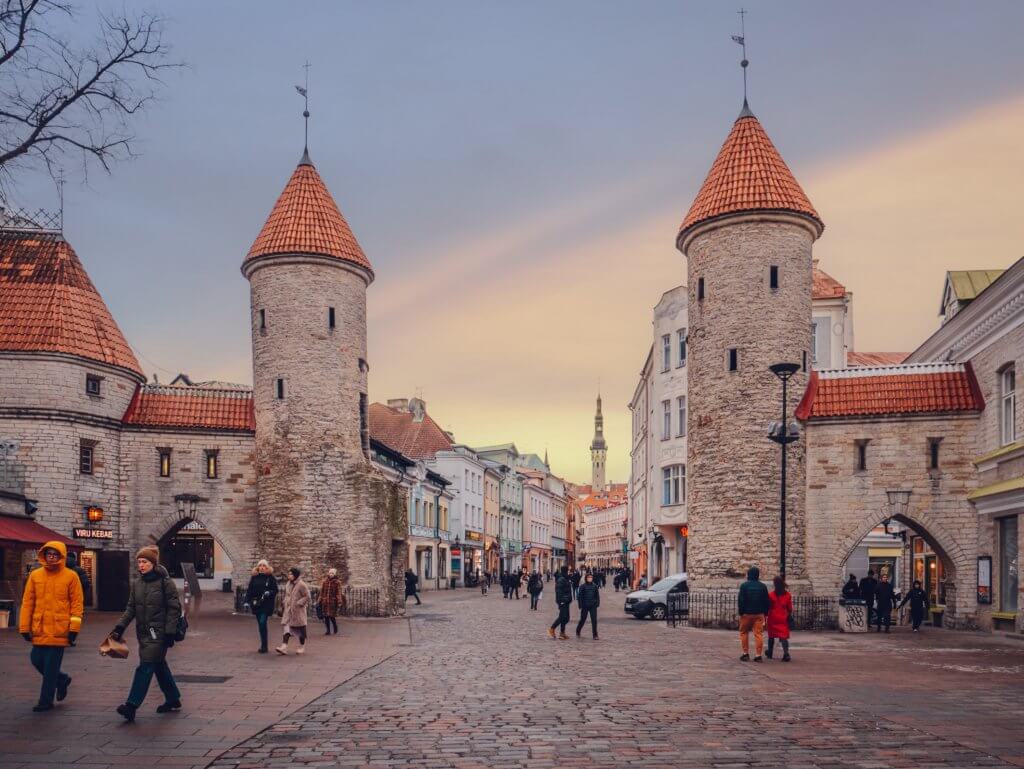 Viru Gate in Old Town Tallinn during sunset. A wide-angle, eye-level shot of the historic Viru Gate in Tallinn, Estonia, at dusk. Two large, cylindrical stone towers with conical terracotta-tiled roofs flank a cobblestone entrance leading into the Old Town. The street is bustling with people in winter clothing, including a person in a bright yellow jacket on the left and a pair walking together on the right.
In the background, a long street lined with pastel-colored buildings recedes toward the slender, distant spire of the Town Hall. The sky is a soft gradient of pale blue and warm orange, with a faint, wispy cloud stretching across the horizon. Shops and restaurants with glowing windows, including a "Viru Kebab" sign, are visible at the base of the stone structures.
