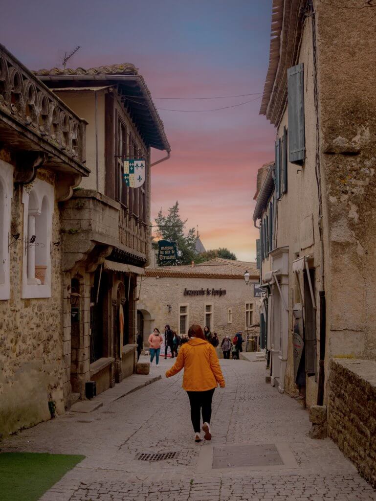 A medium-angle shot from behind shows a person in a bright yellow jacket and black pants walking down a narrow, cobblestone street.

The street is lined with historic stone buildings featuring unique architectural details, such as a decorative stone balcony on the left and various hanging shop signs. In the background, other pedestrians are visible on the slightly downhill path. The scene is set against a dramatic sky with soft orange and purple hues from a sunset, visible just above the rooftops at the end of the alley.
