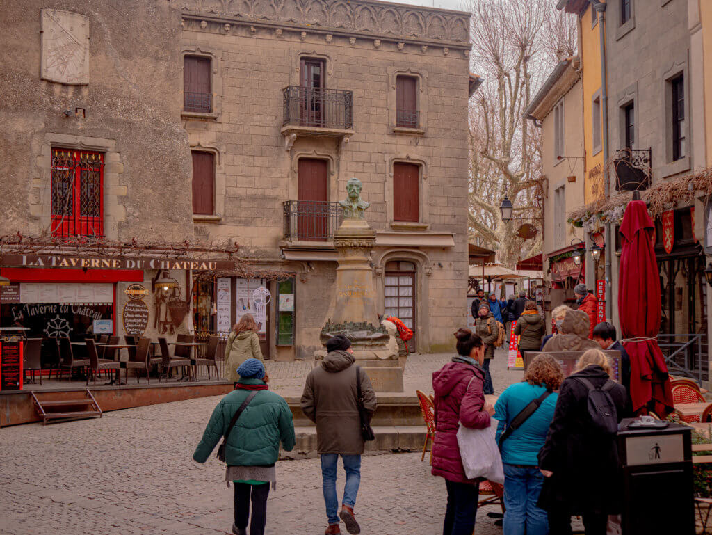 Medieval city of Carcassonne France. A wide, eye-level shot captures a busy cobblestone square in a historic European town. In the center, a bronze bust sits atop a tall, ornate stone pedestal.

Behind the monument stands a weathered, multi-story stone building featuring several windows with dark brown shutters and small wrought-iron balconies. To the left, the ground floor houses a restaurant named "La Taverne du Château," marked by a red storefront and outdoor seating with dark chairs and tables.

Numerous people dressed in winter clothing—including jackets, beanies, and scarves—are walking through the square or standing near the restaurant. In the background, bare trees rise behind the buildings under an overcast, grey sky.