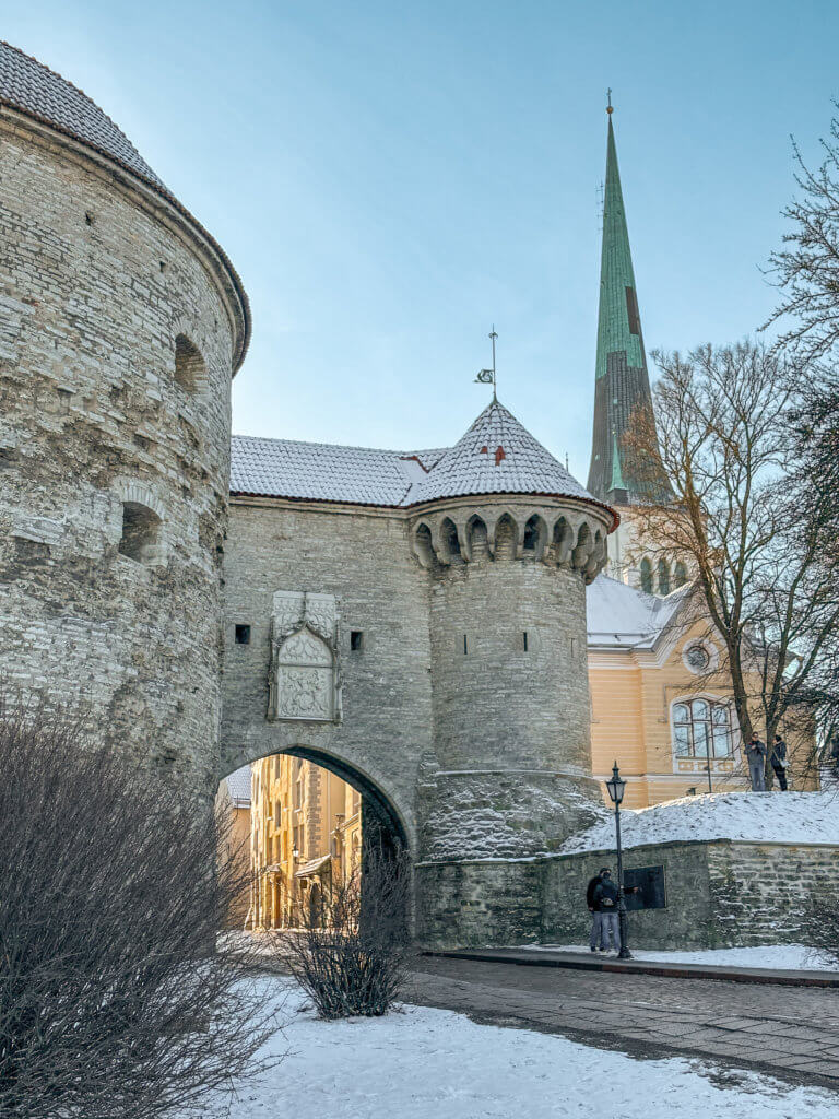 Tallinn in winter. A vertical shot of the historic Great Coastal Gate and Fat Margaret tower in Tallinn, Estonia, under a bright, clear sky. The massive, circular Fat Margaret stone tower stands on the left, connected to a smaller defensive tower by a stone archway that serves as an entrance to the Old Town. A decorative white stone relief is mounted above the arch.
In the background, the tall, slender green spire of St. Olaf’s Church rises high above the surrounding buildings. Patches of white snow cover the ground and the rooftops of the fortifications. A few people are visible near the stone walls on the right side of the image.
