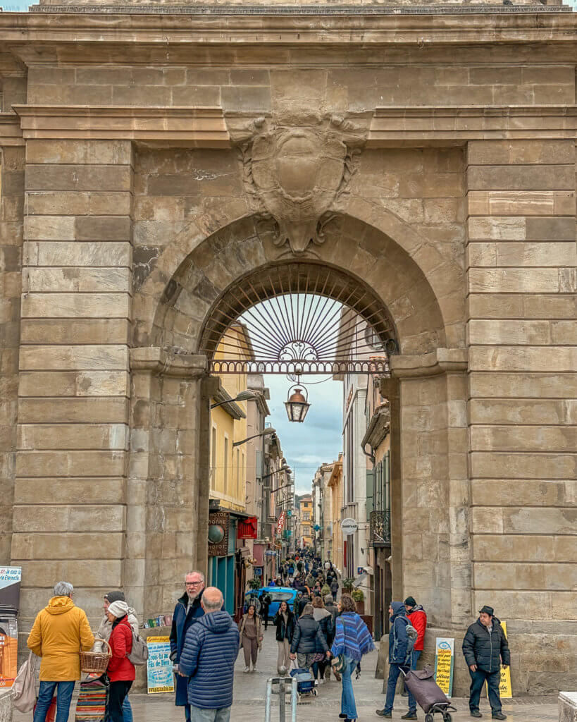 Porte des Jacobins Gate in Carcssonne. A wide, eye-level shot captures a large, historic stone archway with a decorative crest at its peak. Through the arch, a busy, narrow street stretches into the distance, lined with multi-story buildings and various shop signs.

In the foreground and moving through the arch, numerous people are visible, many dressed in winter coats and hats. A man in a bright yellow jacket stands to the left, and several others are walking toward or away from the camera. The street beyond the archway is densely packed with pedestrians, creating a lively urban atmosphere under a cloudy, overcast sky.