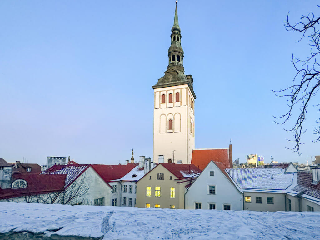 A high-angle, wide shot of the snow-covered rooftops of Tallinn, Estonia, featuring the prominent white tower of St. Nicholas' Church against a clear, pale blue sky. In the foreground, a thick layer of textured white snow sits atop a stone wall, while the midground is filled with traditional buildings with vibrant red-orange tiled roofs and pastel-colored facades. The church tower rises vertically in the center, topped with a distinctive dark green spire. Leafless branches frame the right edge of the image, and a modern city skyline is visible faintly in the distant background.