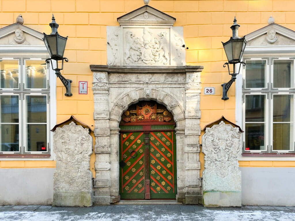 Red green and gold doorway of the house of the brotherhood of blackheads in Tallinn Estonia. A close-up, eye-level shot of a grand, historic entrance in Tallinn, Estonia. The doorway features a heavy wooden double door painted with a diagonal green and reddish-orange diamond pattern, accented by small golden floral studs. It is set within an elaborate arched stone frame with a detailed coat of arms carved into the lintel above.
The door is flanked by two large, weathered stone relief panels featuring intricate heraldic carvings. Two ornate black wrought-iron lanterns are mounted on the pale yellow facade on either side of the entrance. Large, white-trimmed windows are partially visible on the far left and right. The ground in front of the building is paved with grey stone and dusted with a light layer of snow.