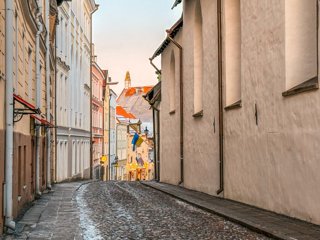 Pastel buildings in Tallinn and cobbled streets covered in snow during winter. A ground-level, slightly upward-angled shot of a narrow cobblestone street in Tallinn, Estonia, receding into the distance. On the right, a long, high white wall of a medieval building features tall, slender arched windows and brown downpipes. To the left, a row of historic pastel-colored buildings in shades of cream, light blue, and pink line the street. In the center background, where the street slopes downward, a blue and yellow Ukrainian flag hangs from a building. The sky above is a pale, soft gradient of orange and blue at dusk, and the cobblestones appear damp with patches of light frost or thin ice.