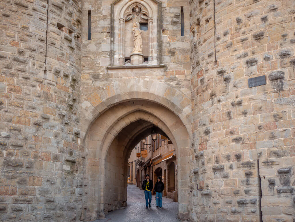 A wide, eye-level shot captures two people walking toward the camera through a massive stone archway in Carcassonne, France.

The archway is integrated into a thick, rugged stone wall made of large, light-colored blocks. Directly above the center of the arch is a decorative Gothic-style niche holding a weathered religious statue. Beyond the gate, a narrow, rising cobblestone street is visible, lined with the warm-toned facades of historic buildings. The sky above the fortifications is a soft, pale blue with light horizontal clouds.