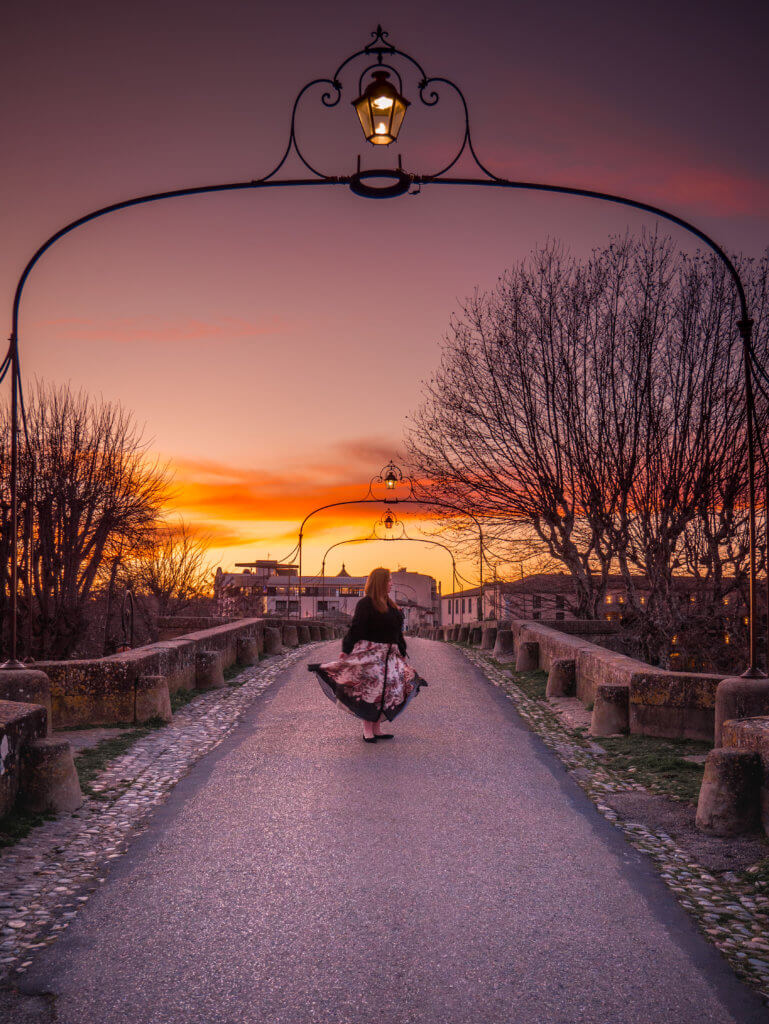 A wide, eye-level shot captures a person in a dark top and a long floral skirt twirling in the center of the Pont Vieux bridge.

The perspective looks down the length of the stone bridge, which is lined with ornate wrought-iron archways, each holding a glowing lantern. The sky is a dramatic sunset with vibrant orange and deep purple tones. Bare trees frame both sides of the bridge in the mid-ground, and the silhouetted rooftops of the lower town are visible in the distance under the warm glow of the horizon.
