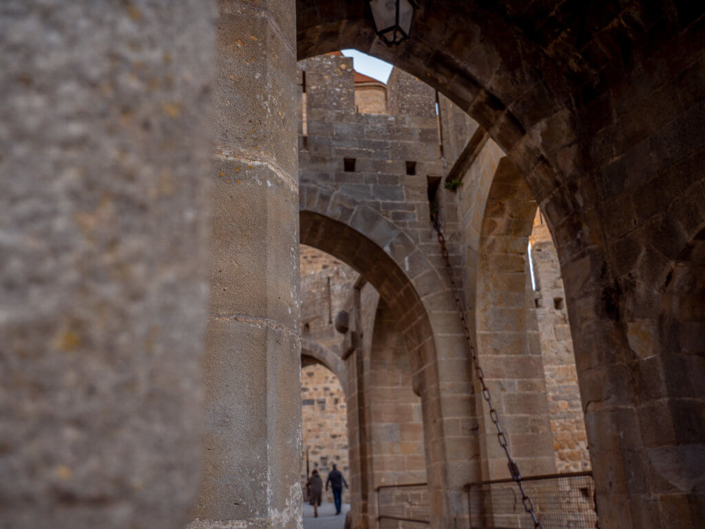 Medieval drawbridge at Carcassonne citadel. A close-up, perspective shot from within the fortifications of Carcassonne, France. The image features a series of massive, weathered stone arches that create a tunnel-like effect.

In the blurred background, two people are seen walking away through the final archway. A heavy metal chain hangs diagonally across the right side of the frame, and a traditional black lantern is visible hanging from the ceiling of one of the arches. The rough, light-brown stonework of the medieval walls dominates the composition, highlighting the scale and age of the fortress.