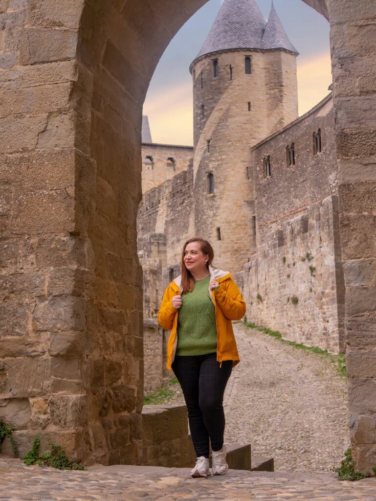 A medium-angle shot captures a person wearing a yellow jacket over a green sweater and black pants, standing on a cobblestone path in Carcassonne, France.

The person is framed by a large, rugged stone archway in the foreground. In the background, the path leads toward the high stone walls of the medieval citadel and a fortified tower with a tall, conical roof. The sky behind the fortifications is a soft blend of orange and pink from a setting sun.