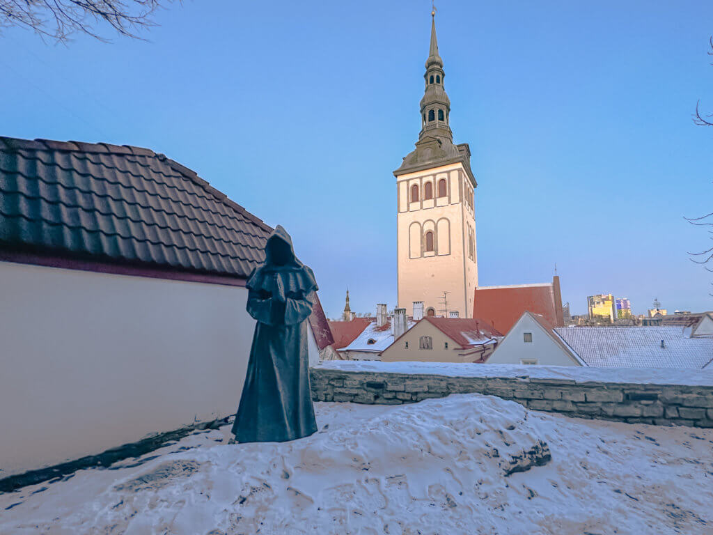 Praying monk statue in the snow at the Danish Kings Garden Tallinn. A medium-angle, wide shot featuring a bronze, faceless monk statue standing on a snow-covered ledge in the foreground. To the right, the prominent white tower of St. Nicholas' Church rises above a cluster of traditional buildings with red-tiled roofs. The rooftops and the ground are blanketed in a layer of bright white snow under a clear, pale blue sky. A white building with a dark, scalloped-tile roof frames the left side of the scene, while a few bare, thin branches are visible on the far right.