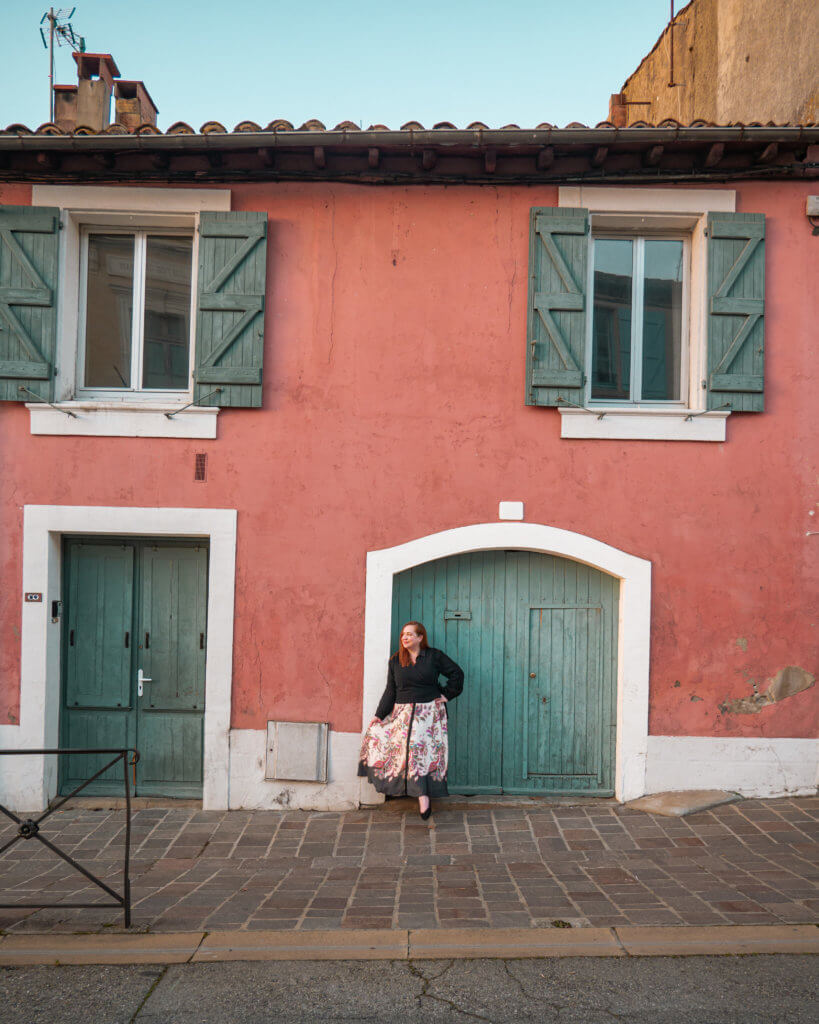 An eye-level, medium shot captures a person with reddish-brown hair standing in front of a weathered, salmon-pink building in Carcassonne, France.

The person is wearing a black long-sleeved top and a flowing white skirt with a bold pink and grey floral pattern. The building features two sets of green wooden shutters on the upper windows and two matching green wooden doors at street level, one of which is set into a white-trimmed archway. The ground is paved with brown stone tiles, and the scene is lit by soft, natural daylight.