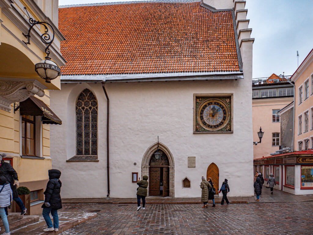 A medium-wide shot of the Church of the Holy Spirit (Pühavaimu kirik) in Tallinn, Estonia. The white, textured facade features an ornate, colorful medieval clock and a large Gothic window with stone tracery. Several people in winter coats are walking across the wet cobblestone square in the foreground, with one person stopping to take a photo of the church. To the left, a yellow building features a decorative hanging sign for "Kohvik Maiasmokk," and to the right, a row of pale buildings and a shop with a red "Money" sign line the street.