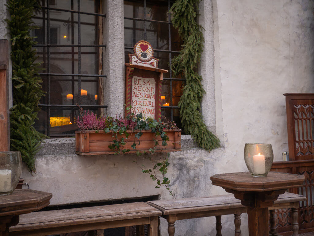 Exterior of Olde Hansa restaurant. A medium shot focuses on the exterior of a medieval-themed restaurant in Tallinn, Estonia. A wooden sign with hand-painted red lettering reads "KESKAEGNE RESTORAN MEDIEVAL RESTAURANT" and is topped with a decorative heart and cauldron illustration.
The sign is mounted on a stone wall between two window panes protected by black iron bars. A wooden window box filled with pink heather and trailing ivy sits below the sign, and thick evergreen garlands frame the window. In the foreground, two rustic wooden tables with lit candles in glass holders are set alongside long wooden benches. Warm candle light is also visible through the window from the interior.