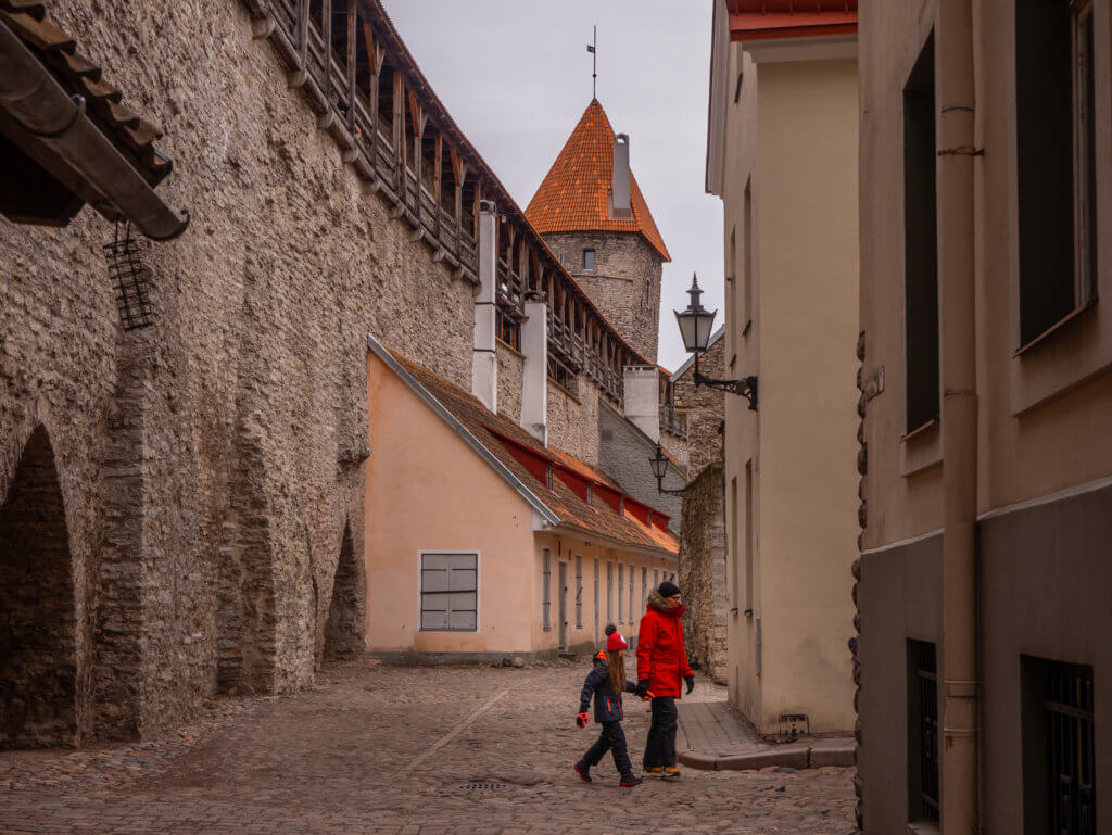 A ground-level view of a historic cobblestone street in Tallinn, Estonia, framed by a massive, rough limestone medieval wall on the left. The high wall features a wooden defensive gallery along its top and deep, arched alcoves at its base.
In the center of the street, two people in winter gear—one in a bright red jacket—walk past a small, single-story peach-colored building built directly against the fortification. In the background, a tall stone defensive tower with a steep, conical orange-tiled roof rises above the walls under a cloudy, overcast sky. Narrow multi-story buildings line the right side of the path, creating a sense of a quiet, enclosed alleyway.