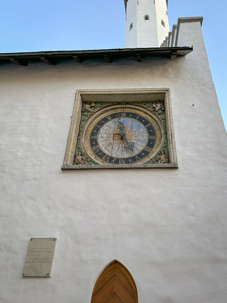 A close-up shot of the ornate, medieval public clock on the white exterior wall of the Holy Spirit Church in Tallinn. The clock face is circular with blue and gold sunburst patterns and Roman numerals, set within a square stone frame decorated with colorful, carved figures in the corners. Below the clock, a small commemorative plaque is mounted on the textured white wall, and the top of a pointed wooden arched door is visible at the bottom of the frame.