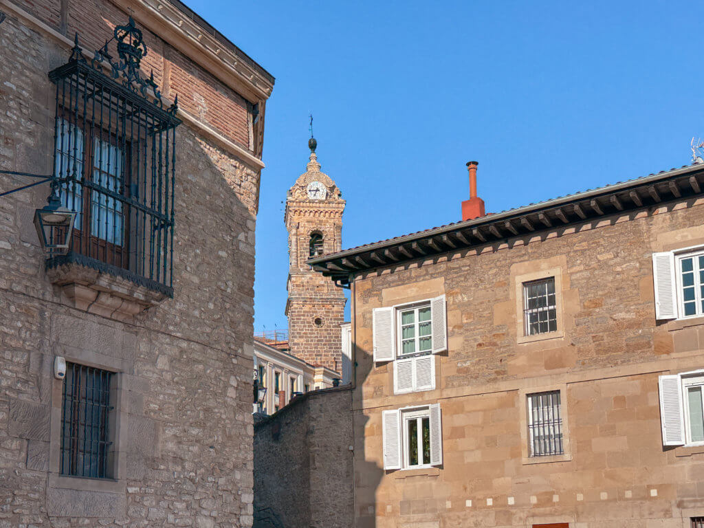 Vitoria Gasteiz A medium shot of a narrow cobblestone street or alleyway in a historic Spanish town, framed by stone buildings on either side. In the center background, an ornate stone clock tower with a bell rises against a clear blue sky.
The building on the left features a decorative black wrought-iron balcony and a traditional street lamp. The building on the right is a multi-story stone structure with white shutters on its windows. The scene is brightly lit by afternoon sun, creating sharp shadows on the stone facades.