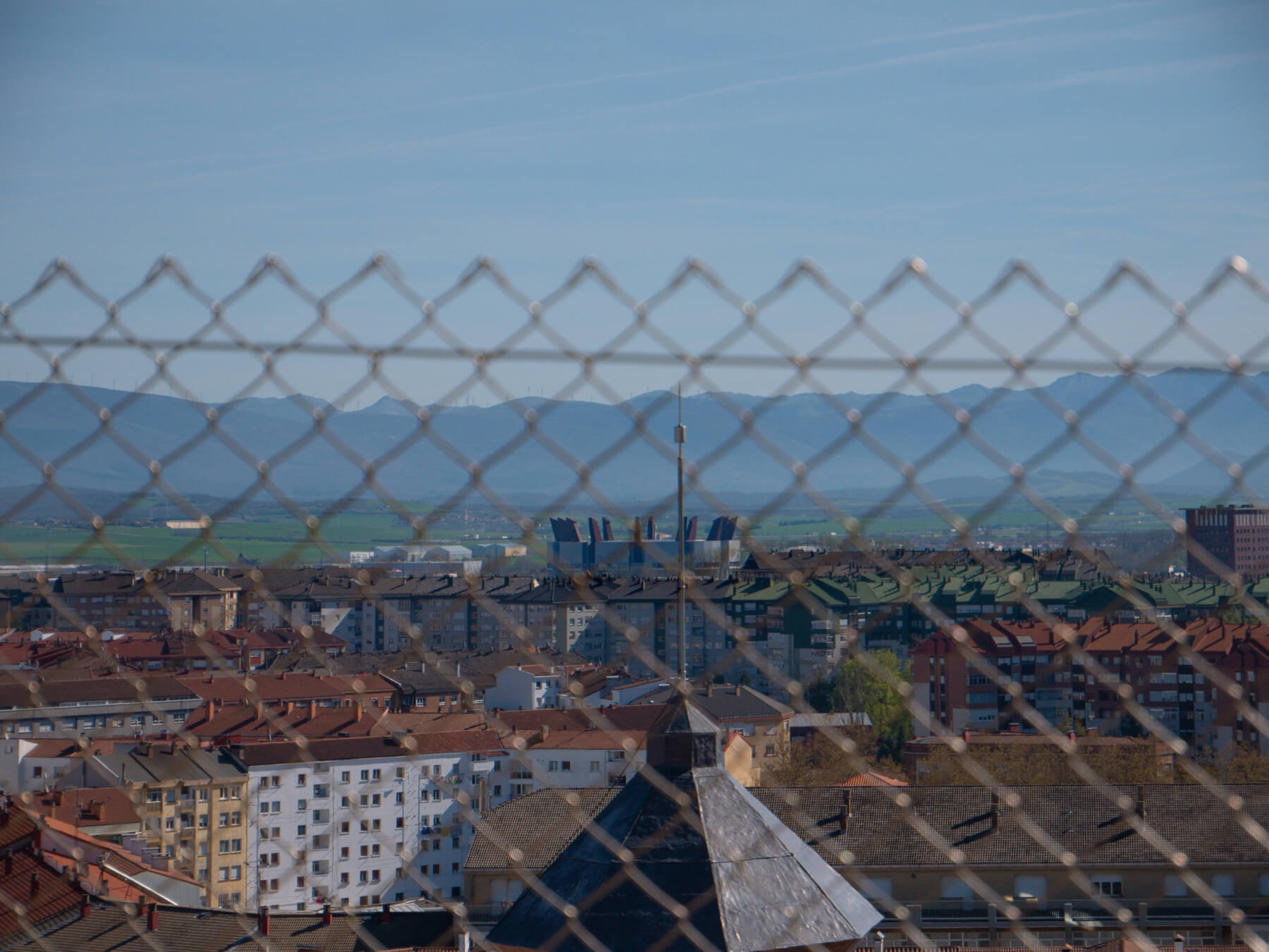An elevated, slightly blurred view of a city's rooftops seen through a chain-link fence in the foreground. The cityscape features numerous multi-story apartment buildings with a mix of red-tiled and flat roofs. In the distance, a range of rolling blue mountains sits under a clear sky. A prominent pointed spire or tower rooftop is visible in the lower center, partially obscured by the metal diamond pattern of the fence.