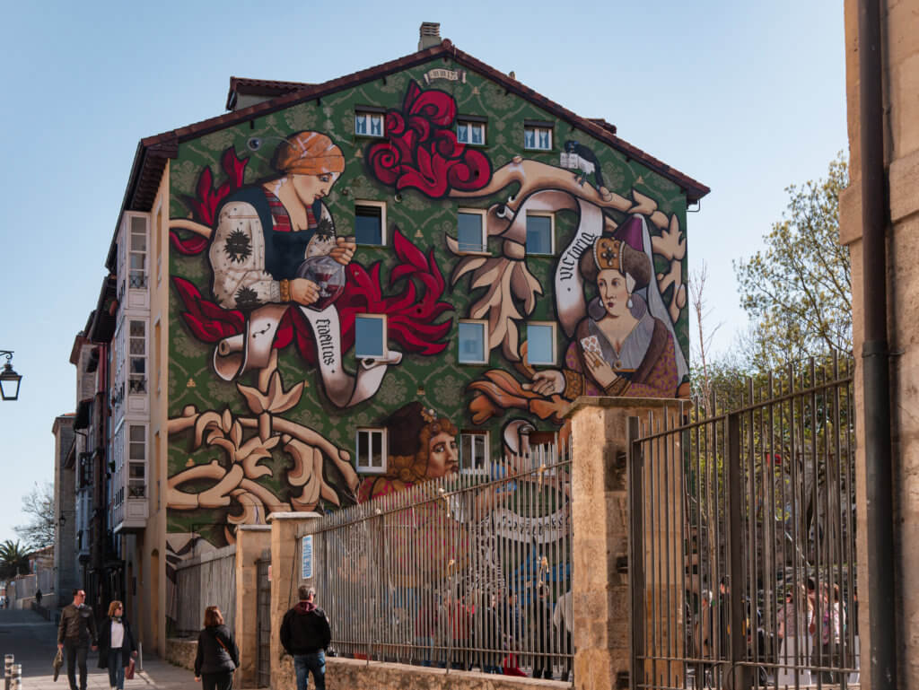 Vitoria Gasteiz An outdoor shot showcases a large, colorful mural painted on the side of a four-story building. The mural, set against a green patterned background, depicts three figures in Renaissance-style clothing and includes flowing ribbons with the Latin words "Fidelitas" and "Victoria."
In the foreground, a metal fence runs along a sidewalk where several pedestrians are walking. The building stands under a clear, pale blue sky, and a portion of a stone structure is visible on the far right.
