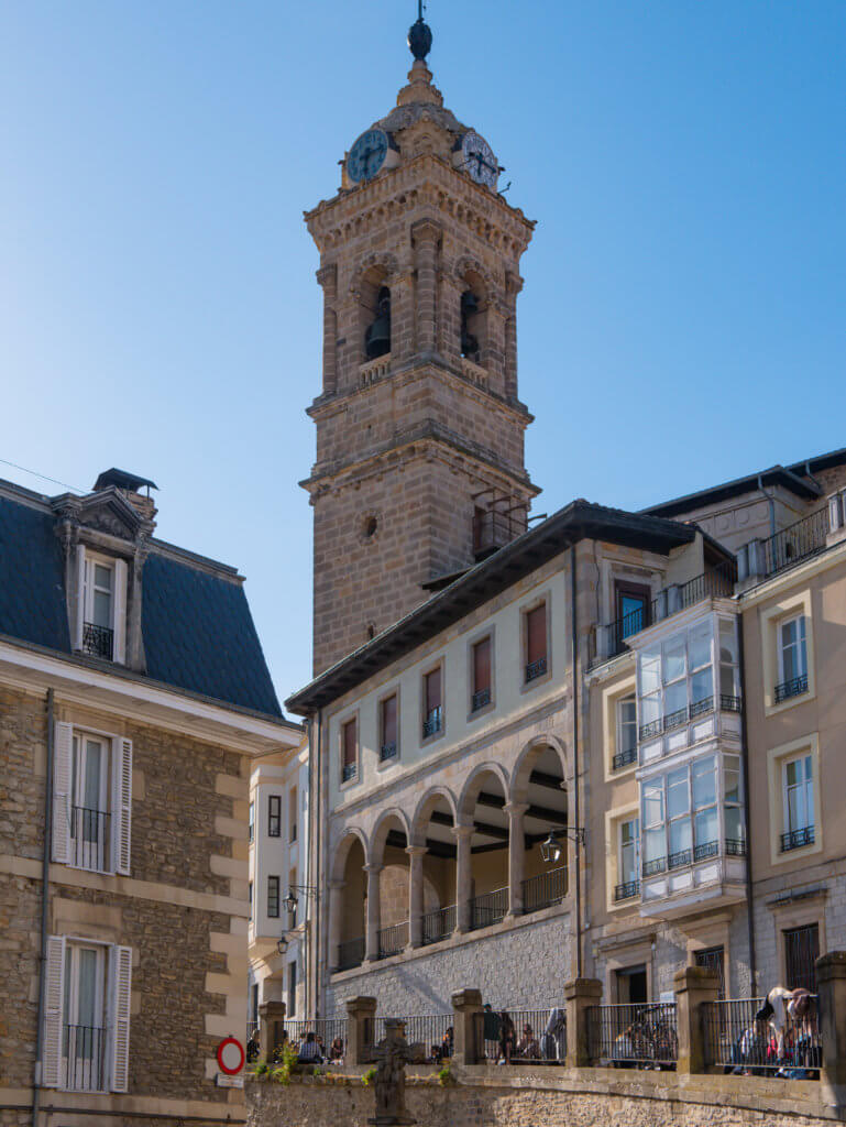 Vitoria-Gasteiz A medium-angle shot captures the ornate stone clock tower of the Church of San Vicente Mártir rising above the surrounding historic architecture in Vitoria-Gasteiz, Spain.
The tower, constructed from light-colored stone, features an arched belfry with visible bells and a decorative top section housing multiple clock faces. To its right, a multi-story building displays a prominent elevated stone gallery with a row of five elegant arches supported by slender columns. In the foreground, a stone retaining wall with a metal railing runs along a lower level where people can be seen sitting in the sun. The scene is set against a clear, deep blue sky, with bright sunlight creating soft shadows across the stone facades.