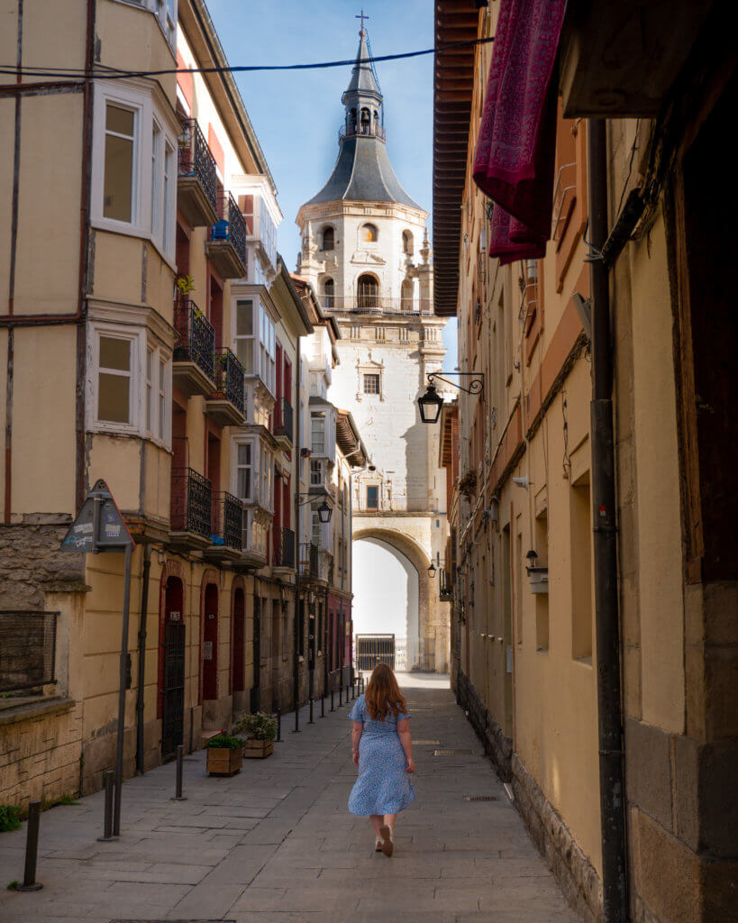 Ahigh-angle view looking down a narrow sunlit stone street in Bilbao's Abando district, where a person in a blue dress is walking toward the towering Curch of San Vicente Martir. The tall historical buildings flanking the lane feature traditional windowed balconiew and warm-toned facades, framing the church's prominent 16th-century bell tower and its large ground-level archway.