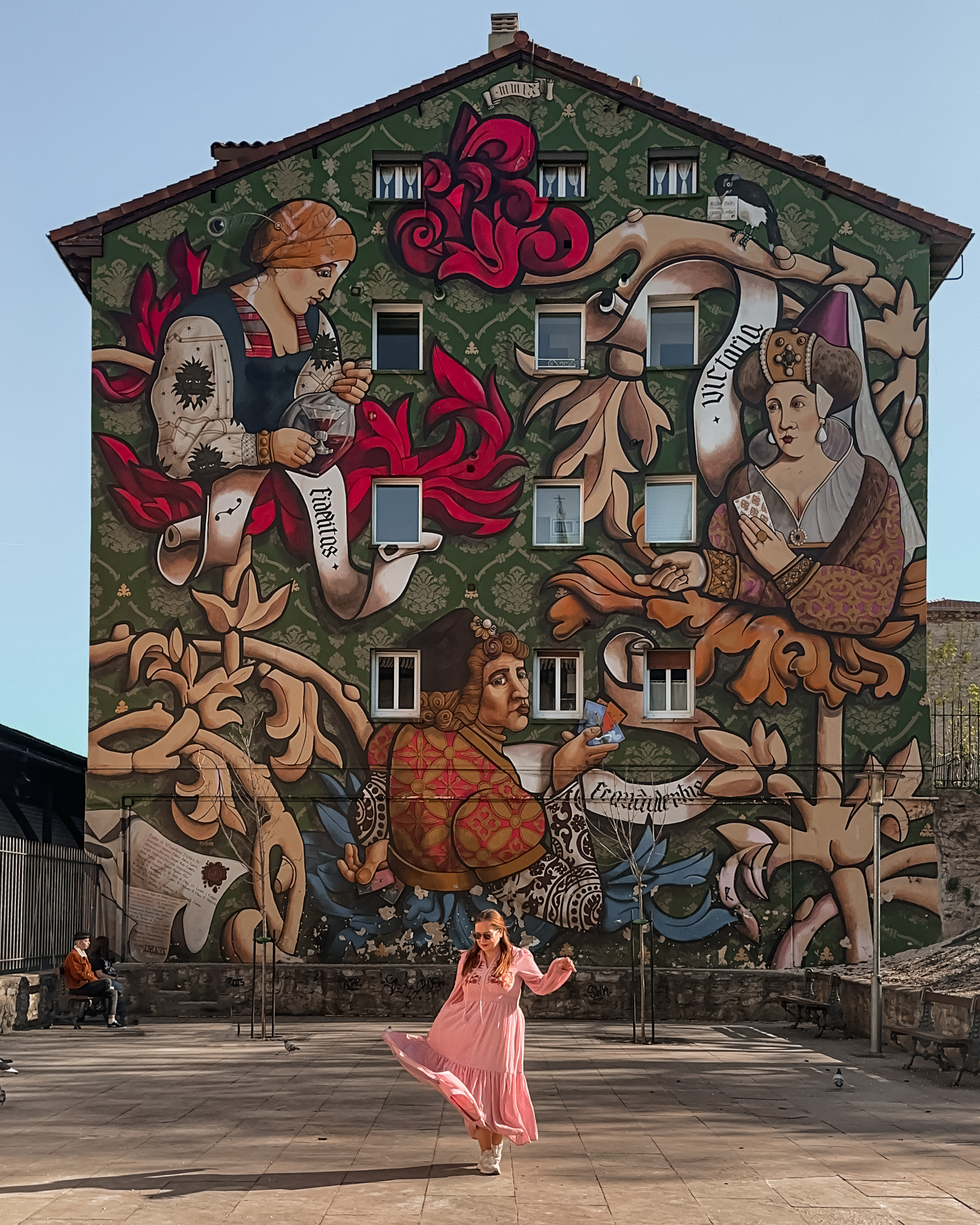 A full, vertical shot captures a person in a flowing pink dress twirling in front of a large, vibrant mural in Vitoria-Gasteiz, Spain. The mural, titled "El Triunfo de Vitoria," covers the entire side of a four-story building and features Renaissance-style figures amidst ornate scrolls with Latin inscriptions like "Fidelitas" and "Victoria" against a patterned green background.
The person is centered in the foreground on a paved stone courtyard, their dress swirling with movement. In the background, to the left, two people sit on a bench near a metal fence, while the scene is illuminated by bright, direct sunlight under a clear sky.