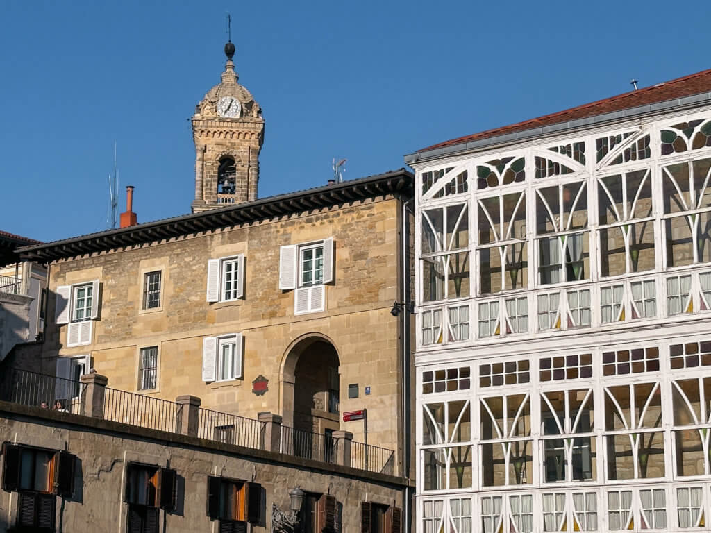Vitoria-Gasteiz An eye-level, medium shot captures the historic architecture of Vitoria-Gasteiz, Spain. In the right foreground, a prominent white building features a traditional Basque facade entirely covered in intricate glass-enclosed balconies, known as miradores.
To the left, a multi-story tan stone building with an arched entryway and windows with white shutters stands behind a stone retaining wall with a black metal railing. In the background, the ornate stone clock tower of the Church of San Miguel Arcángel rises above the rooftops against a clear, deep blue sky.