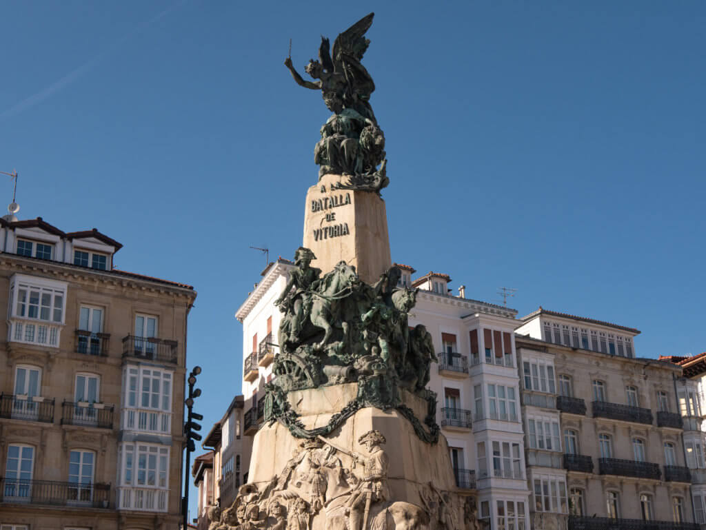 Vitoria-Gasteiz La Batalla de Vitoria monument in Plaza de la Virgen Blanca in Vitoria-Gasteiz. A medium-angle shot captures the Monumento a la Batalla de Vitoria in Vitoria-Gasteiz, Spain. The towering stone monument is adorned with intricate bronze and stone sculptures, including soldiers on horseback and an angelic figure with outstretched wings at the very top. The words "A LA BATALLA DE VITORIA" are inscribed prominently in the center. Behind the monument, several multi-story white and tan buildings with traditional balconies stand against a clear, deep blue sky under bright, direct sunlight.