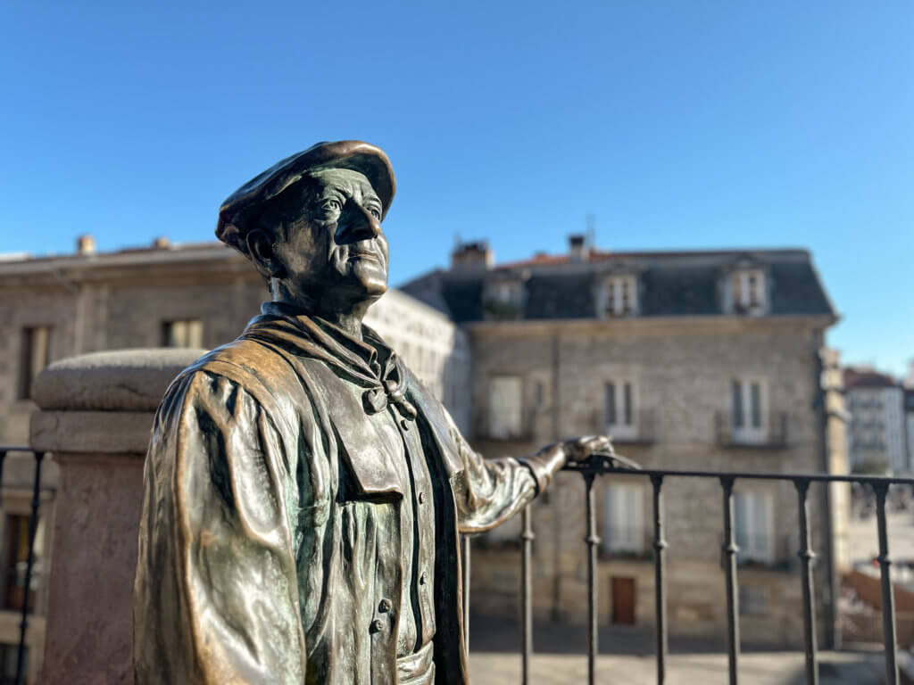 Vitoria Gasteiz A close-up, medium shot of the bronze statue of Celedón, a beloved cultural figure, in Vitoria-Gasteiz. The statue depicts a man with a weathered expression wearing a traditional flat cap and a jacket, standing behind a dark metal railing. The background features the blurred stone facades of historic buildings under a clear, bright blue sky, with sunlight highlighting the metallic texture and detailed folds of the statue's clothing.