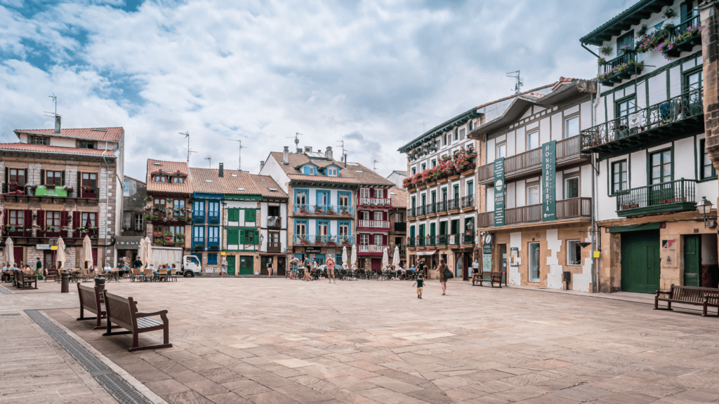 A wide-angle, daylight shot of Arma Plaza in Hondarribia, Spain, featuring a large, open cobblestone square surrounded by traditional Basque architecture. The multi-story buildings are characterized by colorful wooden balconies in shades of blue, green, and red, with some facades adorned with vibrant flower boxes. In the background, people sit at outdoor cafe tables under white umbrellas, while a few pedestrians and a small child walk across the expansive stone plaza. The sky above is bright with soft, scattered white clouds.