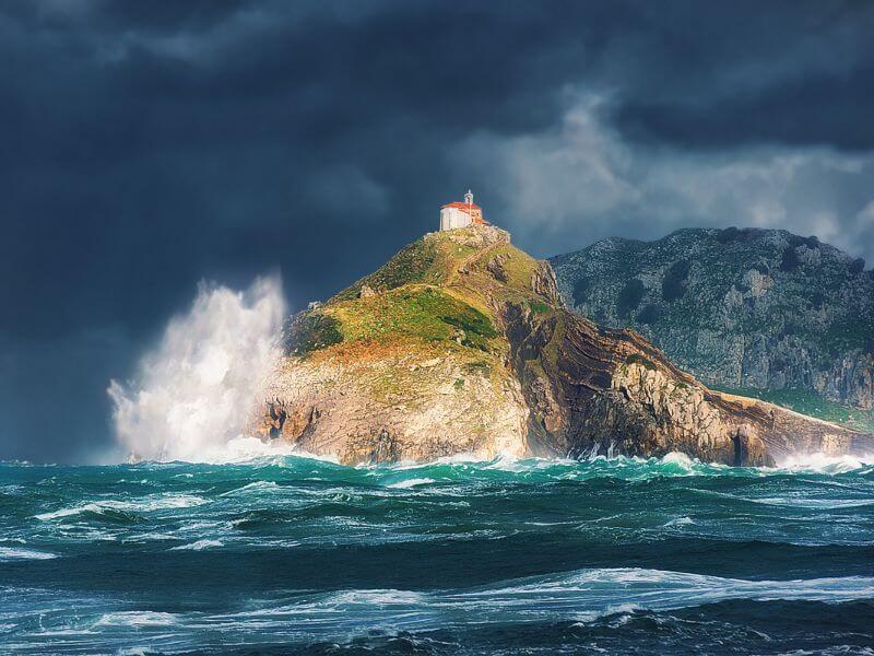 A dramatic view of San Juan de Gaztelugatxe, a rocky islet on the Biscayan coast of Spain, as a mssive white wave crashes against its base. The islet is topped by its historic 10th-century hermitage and surrounded by stormy turquoise waters of the Bay of Biscay, all set under a dark, heavy sky with distant green mountaintains in the background.