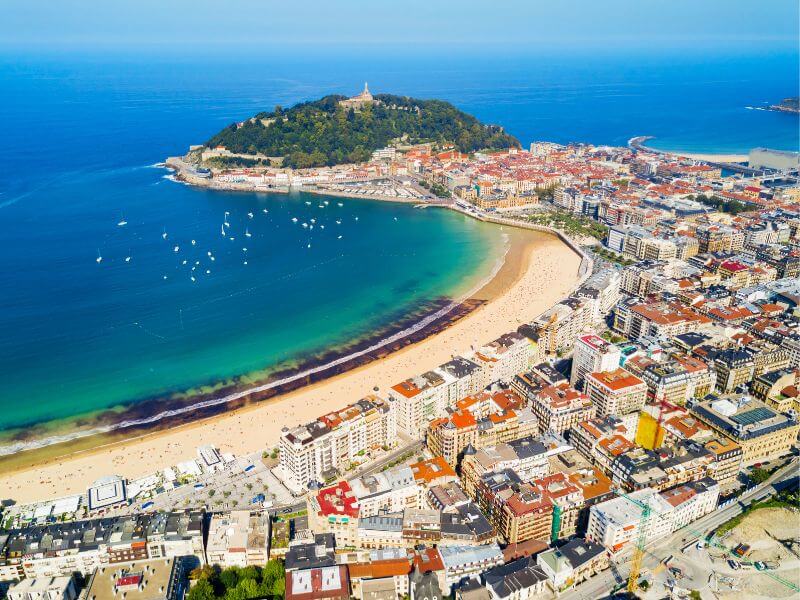 Aerial view of La Concha Beach in San Sebastian, featuring turquoise water with white boats and luxury buildings overlooking the bay.