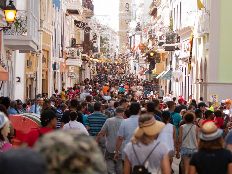 A vibrant and crowded narrow street in San Sebastian's historic Old Town (Parte Vieja), Spain. Thousands of tourists and locals fill the cobblestone lane, which is flanked by tall, colourful traditional buildings featuring ornate balconie and white facades. The street is bustling with social life, typical of the city's famous pintxo bar and shopping district. In the distance, the ornate bell tower of the Basilica of Santa Maria del Coro or the San Sebastian Cathedral rises above the rooftops. Warm sunlight filters down between the buildings, illuminating the dense crowd as they stroll through the heart of the Basque Country's culinary and cultural centre.