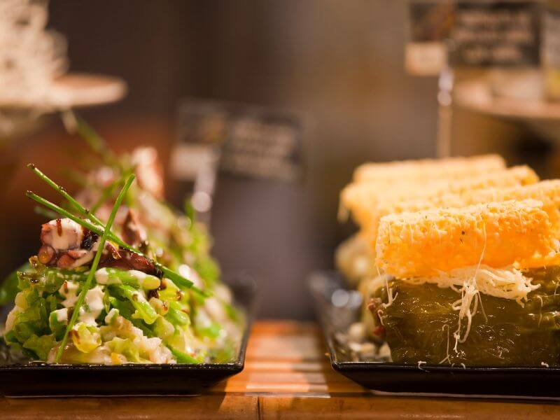 A close-up, shallow focus shot of two distinct Basque pintxos (miniature appetizers) displayed on a bar counter in San Sebastian.