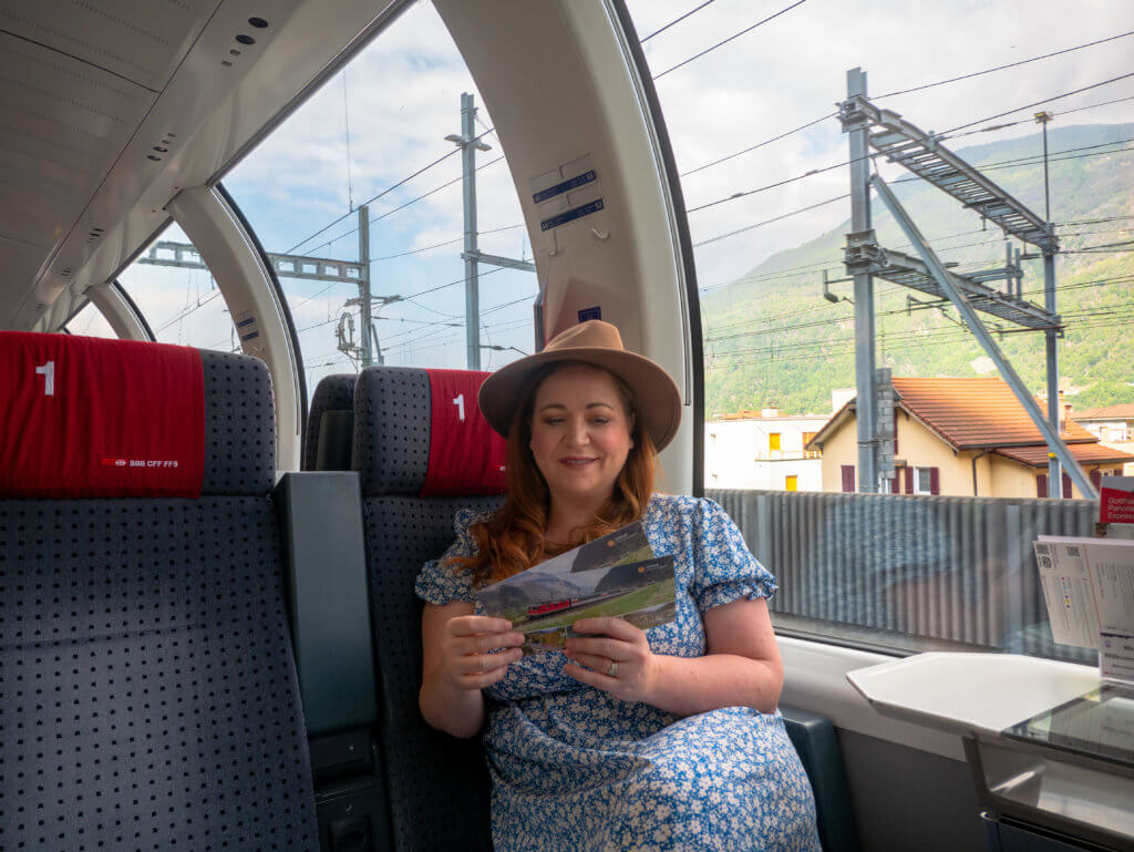 Nicola Lavin, travel writer with Our Unique Stays, wears a hat and a blue dress and reads postcards on the Gotthard Panorama Express