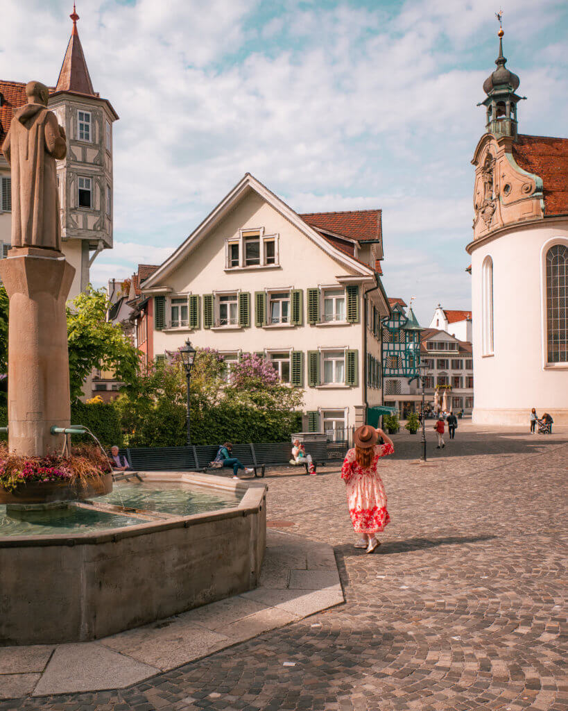 Nicola Lavin, a travel writer with Our Unique Stays, is wearing a floral dress and a fedora hat exploring the fairytale Swiss city of St. Gallen Old Town