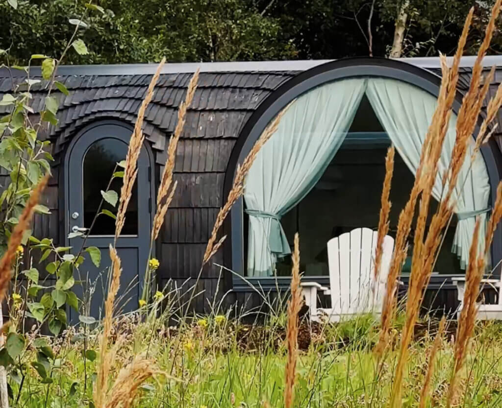 Luxury cabin in Ireland surrounded by wild grasses and trees, featuring a curved window with light blue curtains and a duck-egg Adirondack chair on the porch—a tranquil and unique stay in nature.