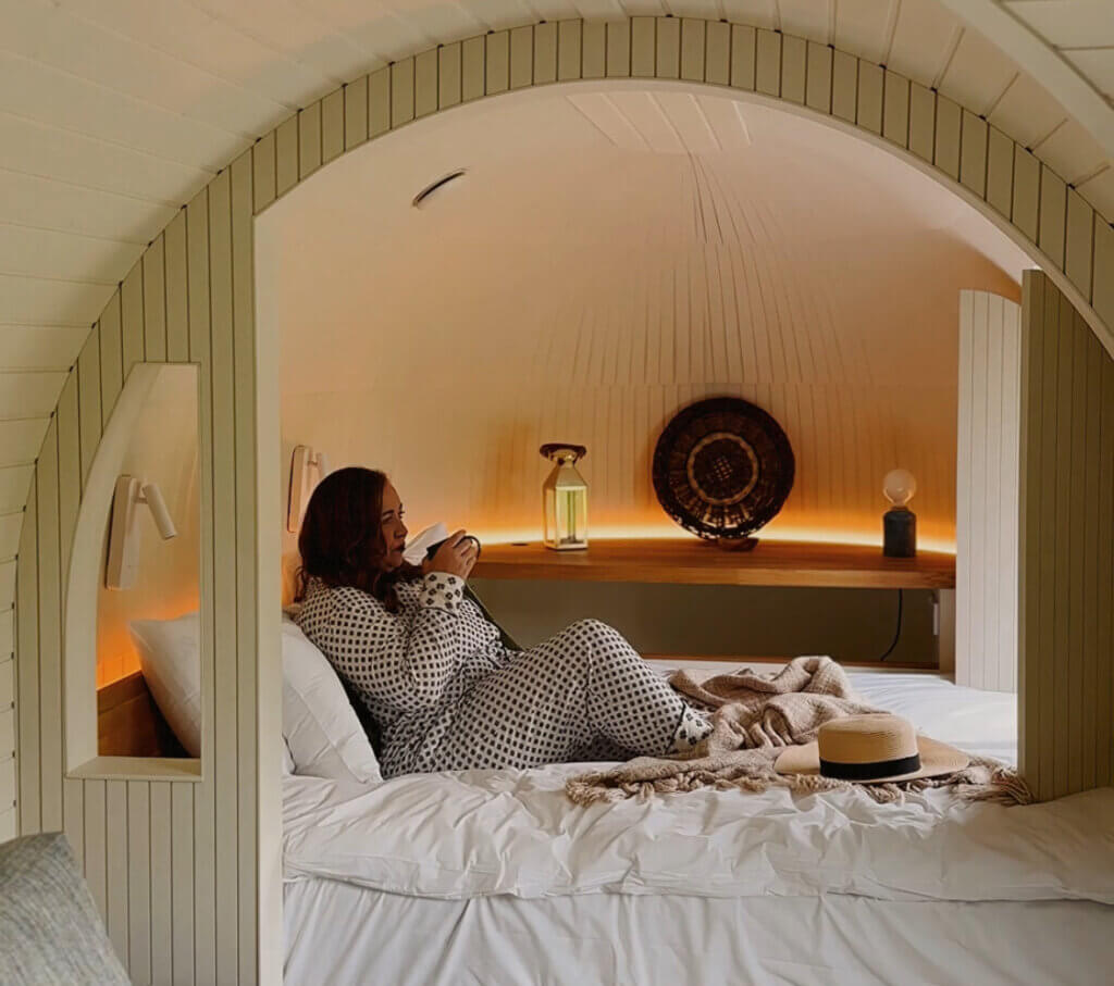 A woman sips coffee in bed in a luxury cabin in Ireland. The interiors are warm and cosy.