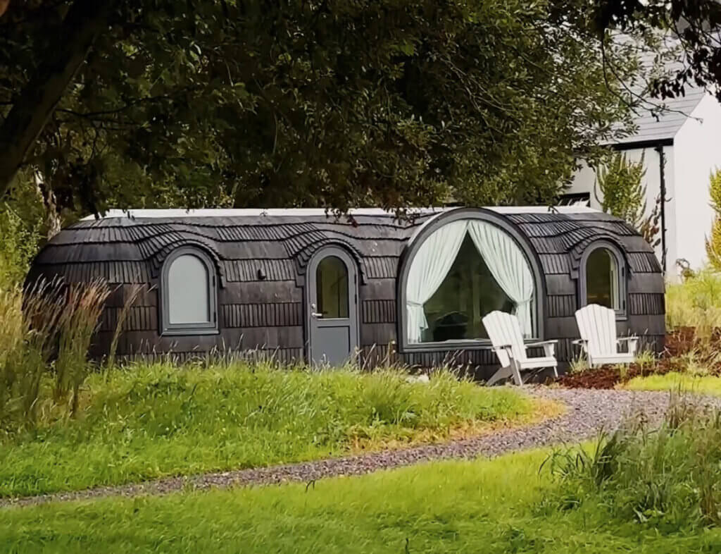 Unique luxury cabin in Ireland with curved architecture, shingle cladding, large round windows, and duck-egg Adirondack chairs set along a gravel path in a lush green landscape.
