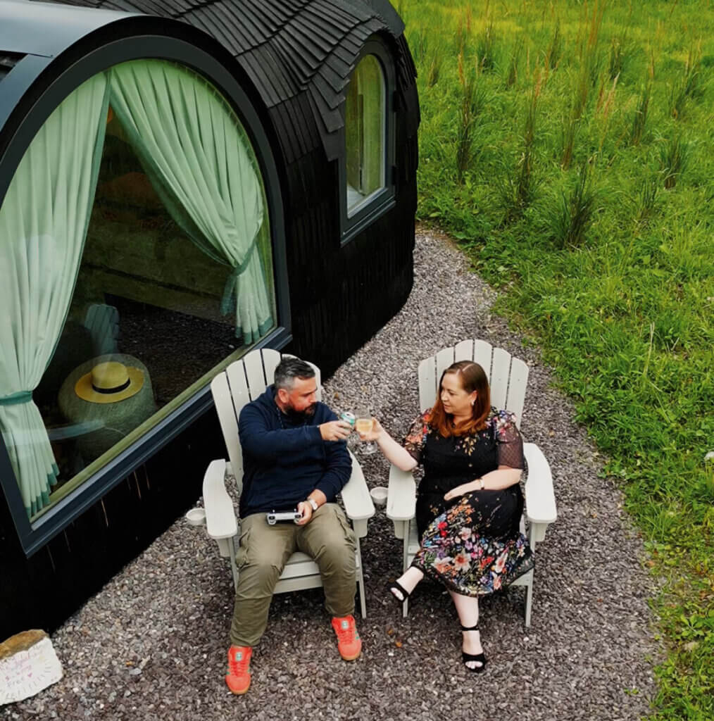 Couple enjoying drinks outside a luxury cabin in Ireland, sitting on duck-egg Adirondack chairs beside a uniquely designed pod-style cabin with curved architecture, set in a lush green countryside.