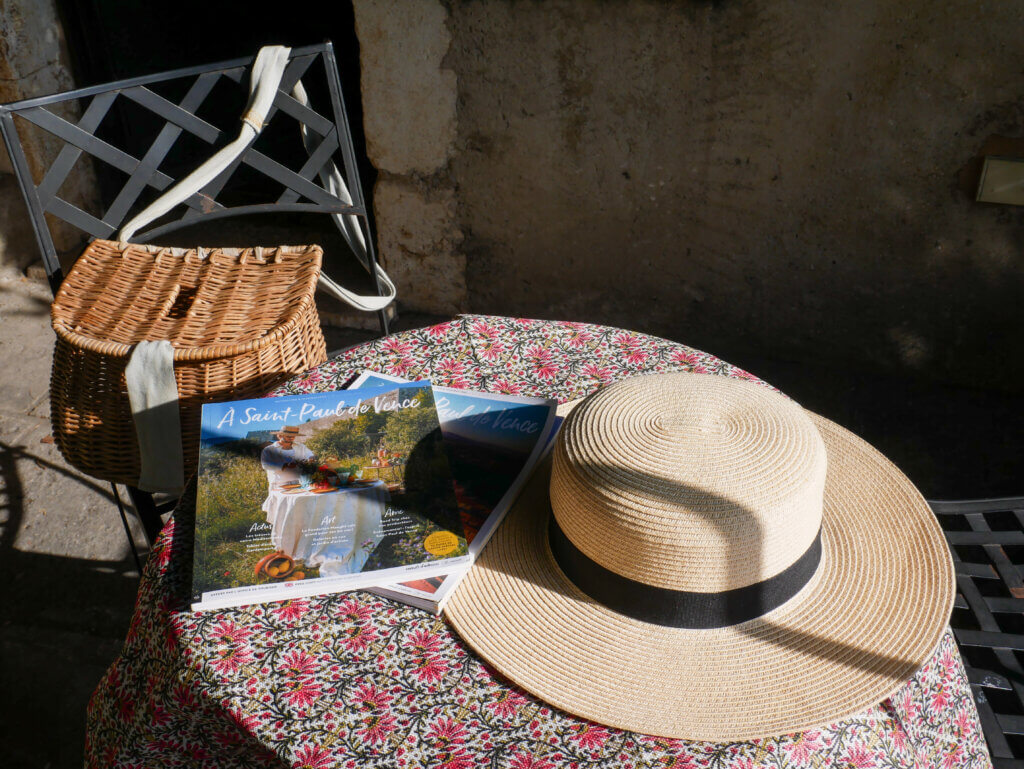Rustic table in the front porch of La Miette. On the table are a straw hat and travel guides on Saint Paul de Vence. On the chair is a picnic basket.