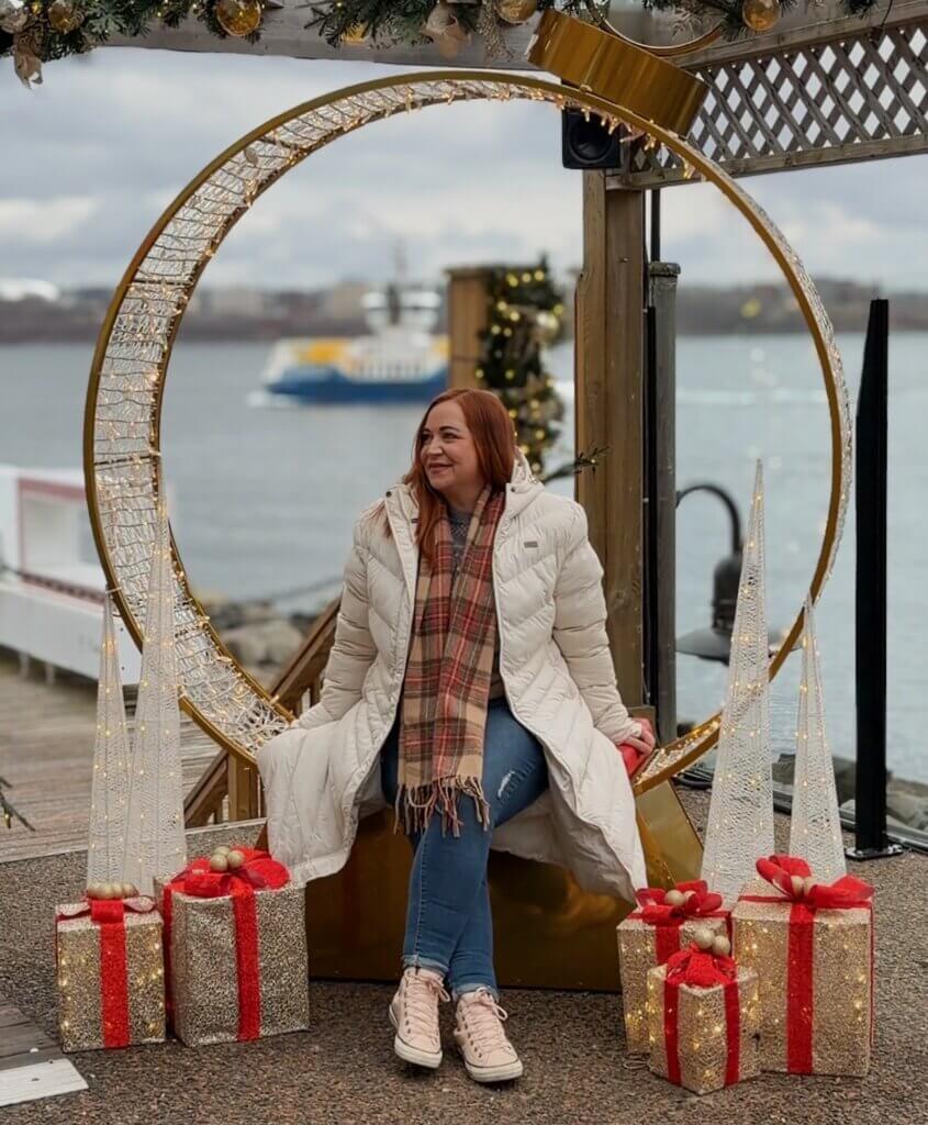 Woman sits on a Christmas display on Halifax harbour for a photograph. She wears a white coat and a tartan scarf.