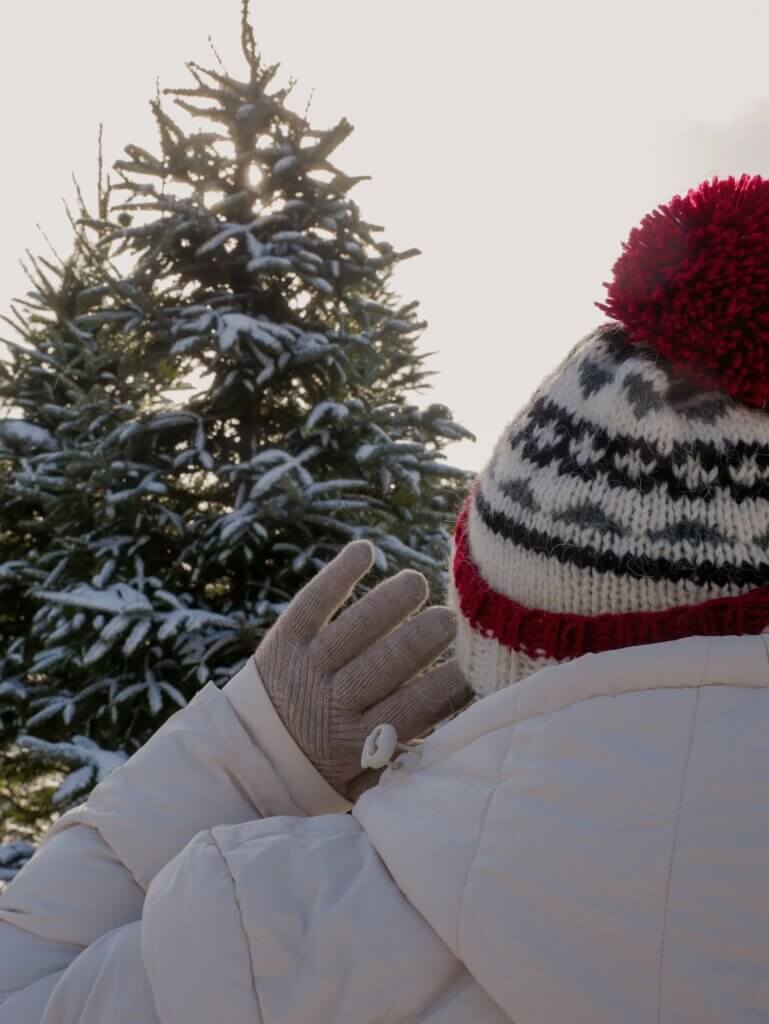 Woman in a white coat and wearing a winter hat and gloves looking at a snowy Christmas tree in Nova Scotia at Christmas.