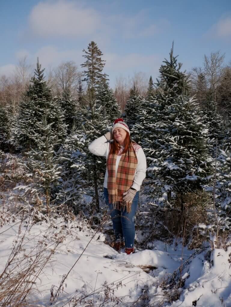 Woman wearing a Christmas jumper and scarf is standing beside snow covered Christmas trees on a Christmas tree farm in Nova Scotia.