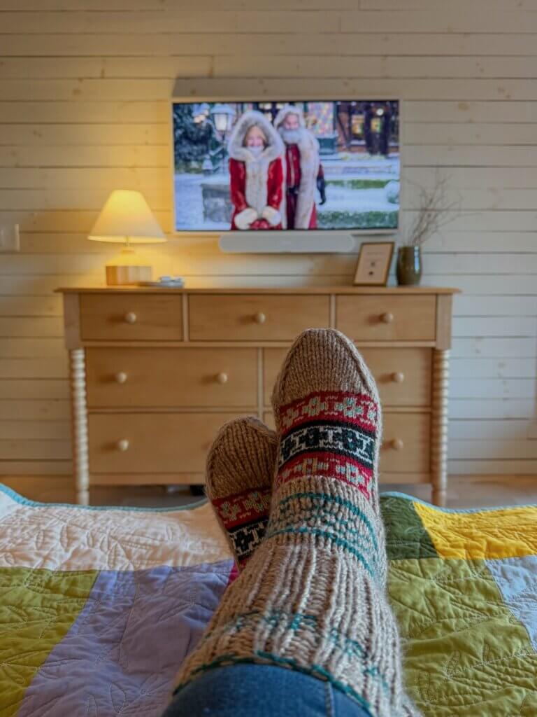 Woman wearing cosy knitted socks rests on a bed watching Christmas movies.